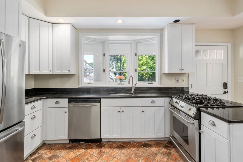 Kitchen with white cabinets, black countertops, stainless steel appliances, and a sink beneath three windows.