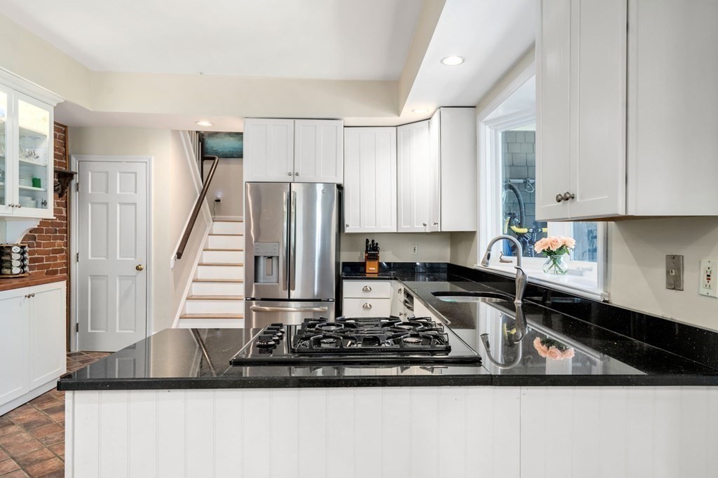 Kitchen with black stone counters, a gas cooktop, stainless refrigerator, and stairs beside the cabinetry.