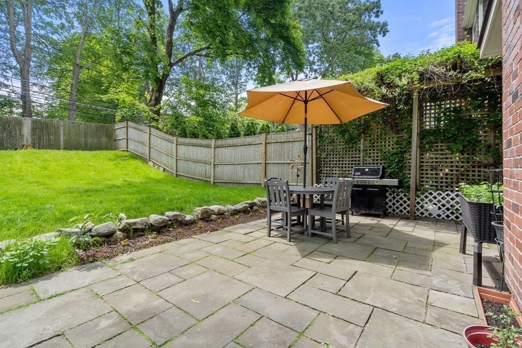Backyard patio with stone paving, outdoor dining set, tan umbrella, barbecue grill, and fenced lawn.