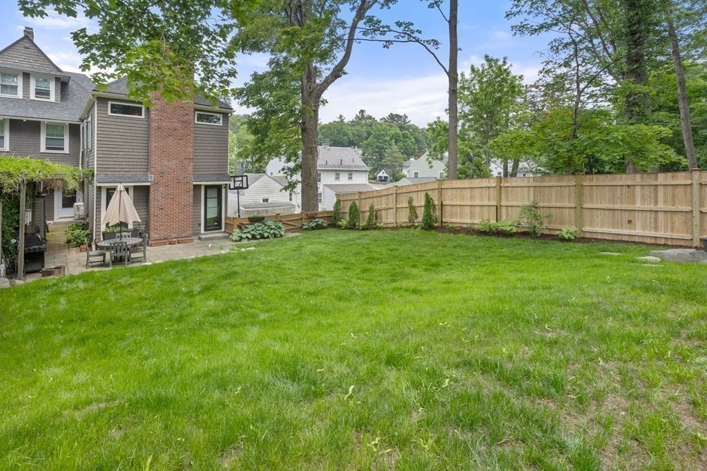 Fenced backyard with a broad lawn, mature trees, and a patio beside the home.