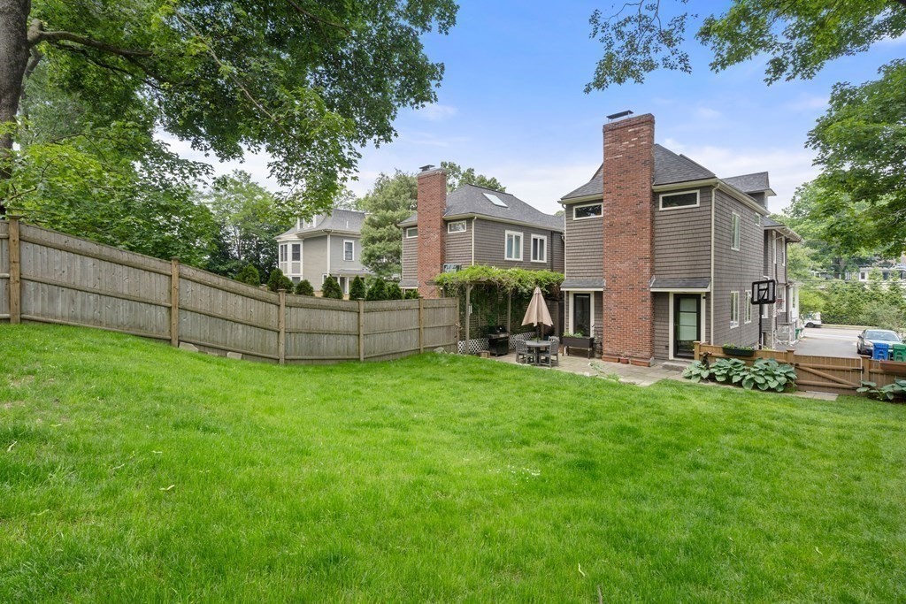 Two-story shingle-sided home behind a broad lawn, with brick chimneys, fenced yard, and patio.