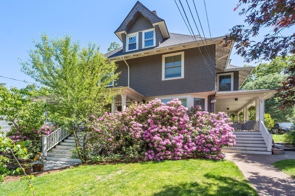 Gray shingle-style house with a covered porch, white stairs, and pink flowering shrubs across the front.