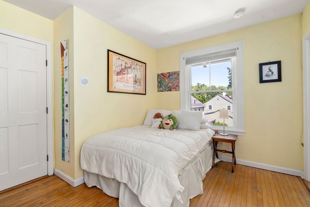 Bedroom with a white bed, yellow walls, hardwood floors, and a window overlooking neighboring houses.