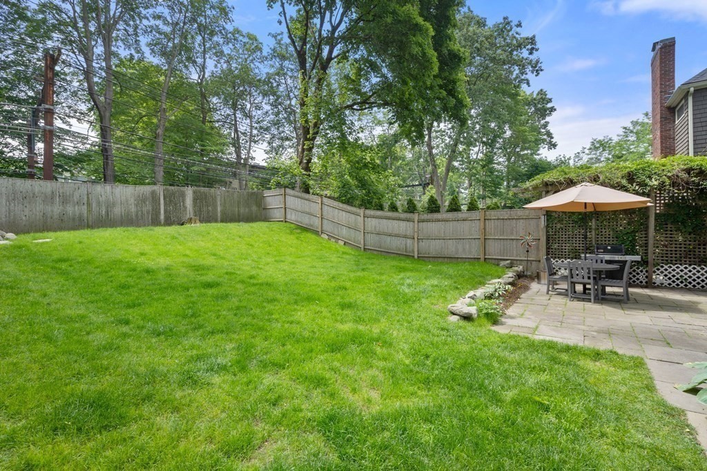 Fenced backyard with a broad lawn and a stone patio dining area beneath an umbrella.