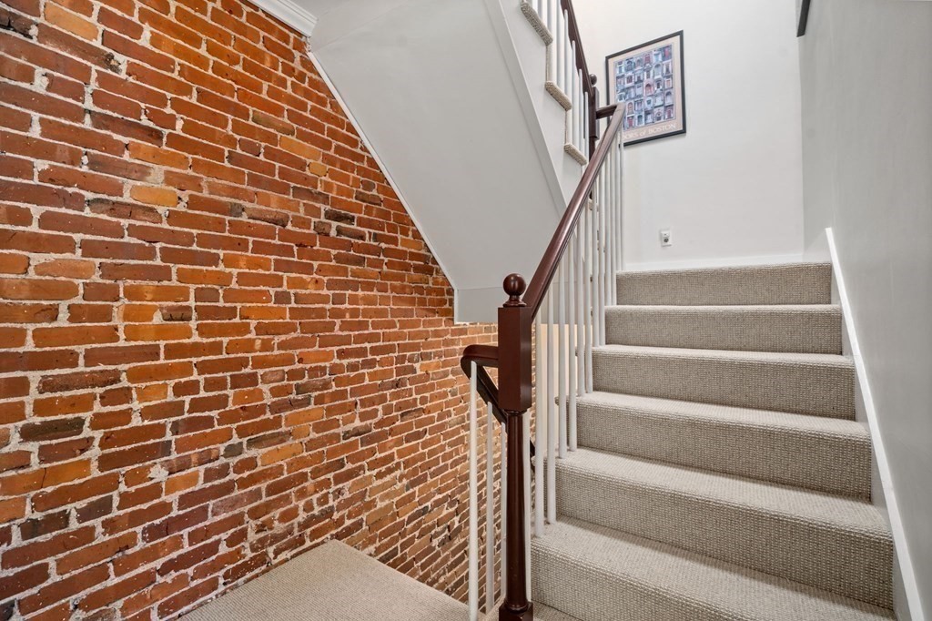 Carpeted staircase with a dark wood railing beside an exposed brick wall.