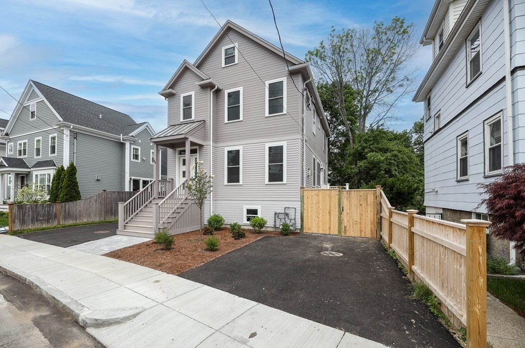 Gray multi-story house with a raised porch, front stairs, wooden fence, and asphalt driveway.