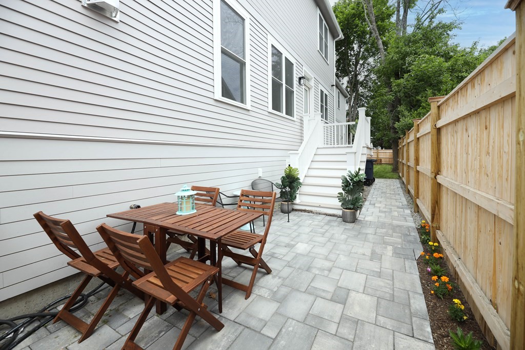 Paved patio beside a white-sided house, with wooden dining set, rear steps, and flowers along a fence.