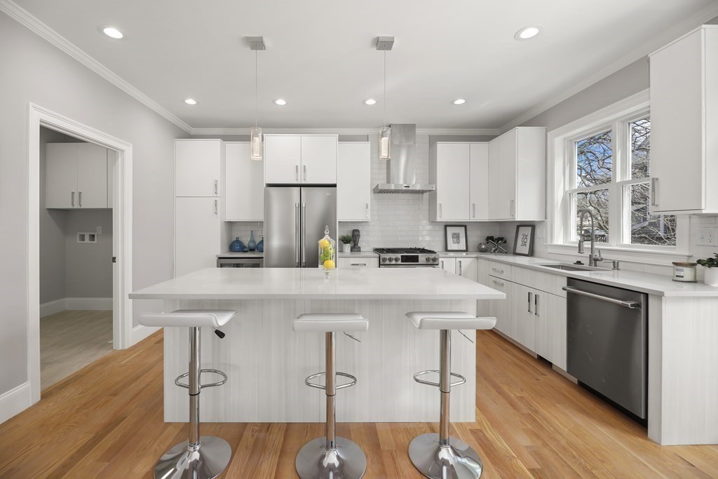 Bright white kitchen with a large island, three bar stools, stainless steel appliances, and wood floors.