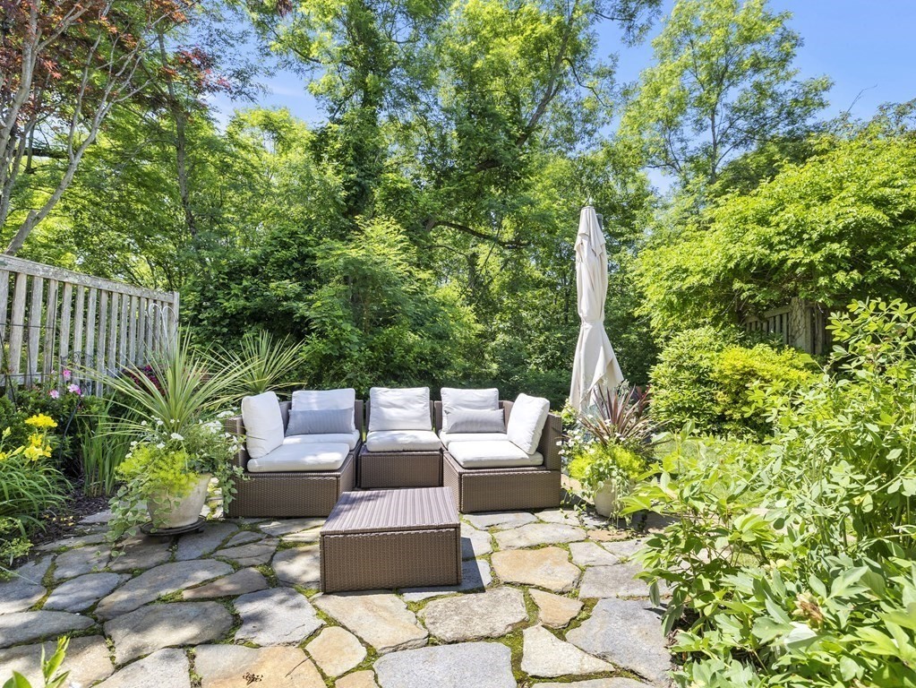 Stone patio with cushioned wicker seating and a closed umbrella, surrounded by lush garden foliage.