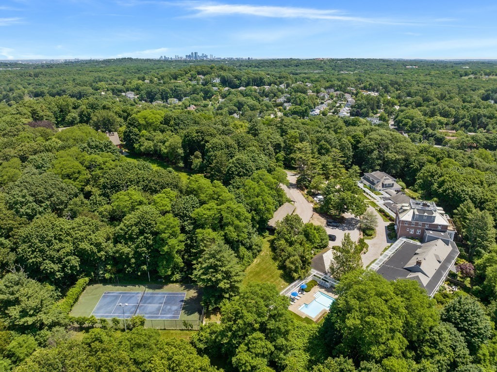 Aerial view of a wooded neighborhood with a pool, tennis courts, and distant city skyline.