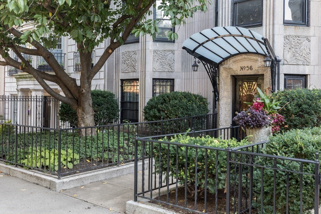 Apartment building entrance beneath a curved glass canopy, framed by a mature tree and landscaped shrubs.