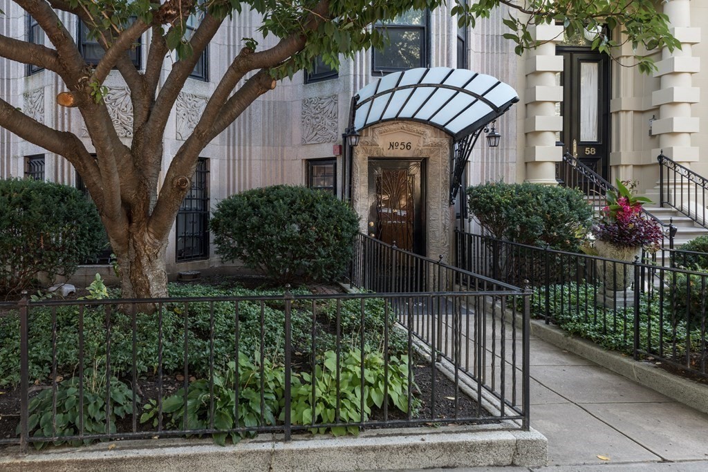 Apartment building entrance with a glass canopy, ornate doorway, and landscaped garden.