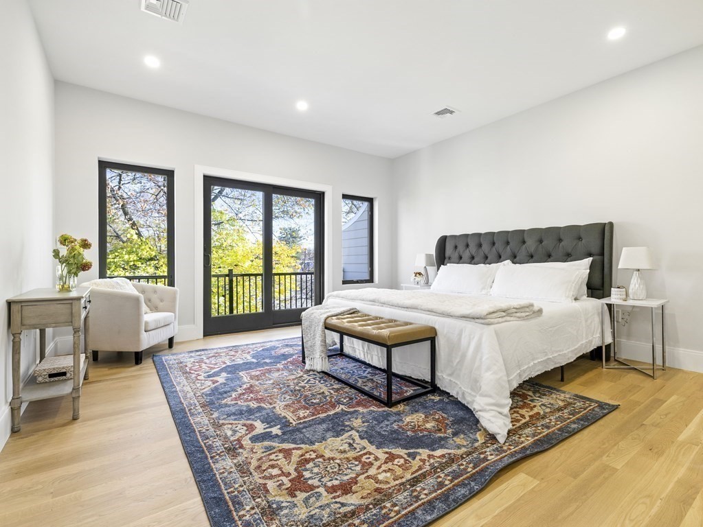 Bedroom with a gray tufted headboard, patterned rug, and balcony doors overlooking leafy trees.
