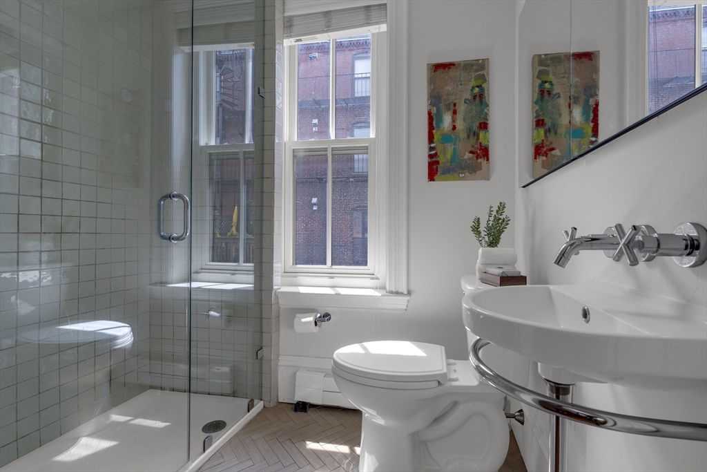 Bright bathroom with a glass shower, pedestal sink, and tall windows overlooking brick buildings.