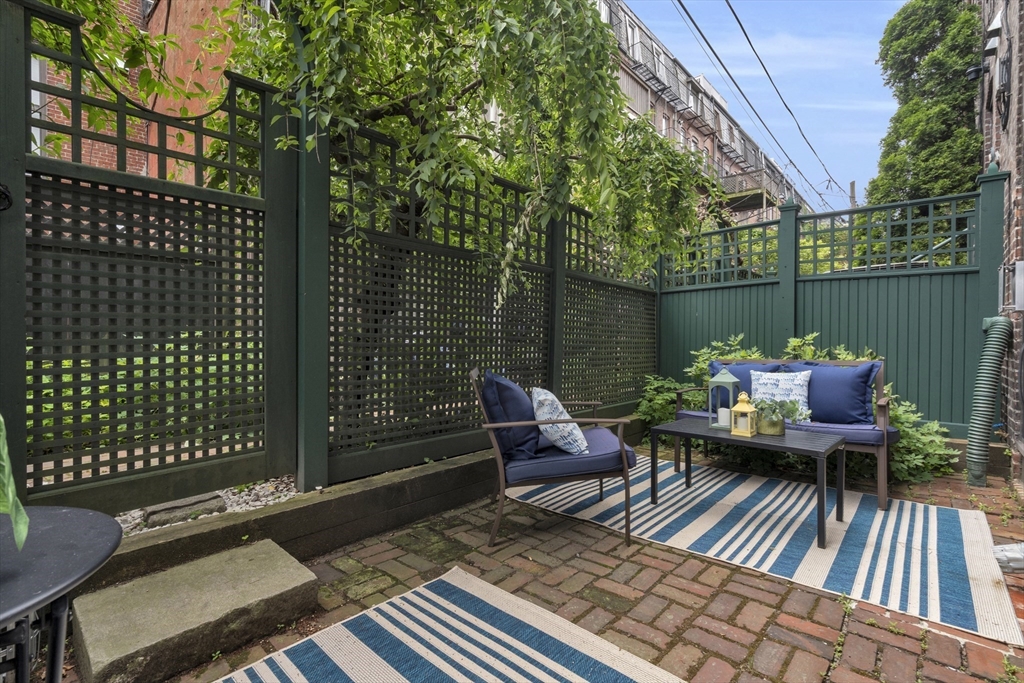 Brick patio with outdoor seating, striped rugs, and a tall green privacy fence.