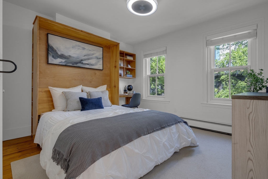 Bedroom with a built-in wood headboard and shelving, a gray bedspread, and two windows overlooking trees.