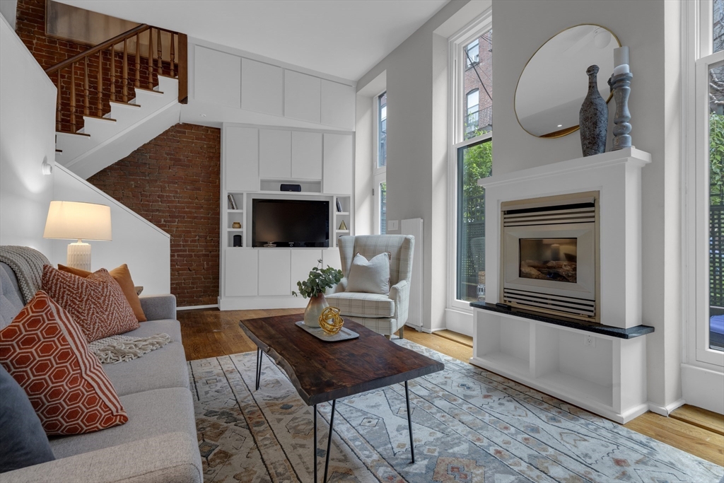Living room with a gas fireplace, exposed brick, loft staircase, and tall windows.
