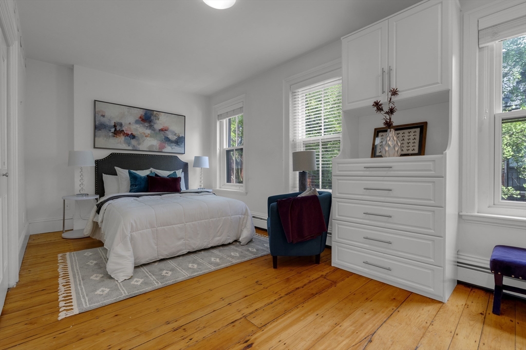 Bedroom with a white bed, built-in dresser and cabinets, teal chair, and wood floors.