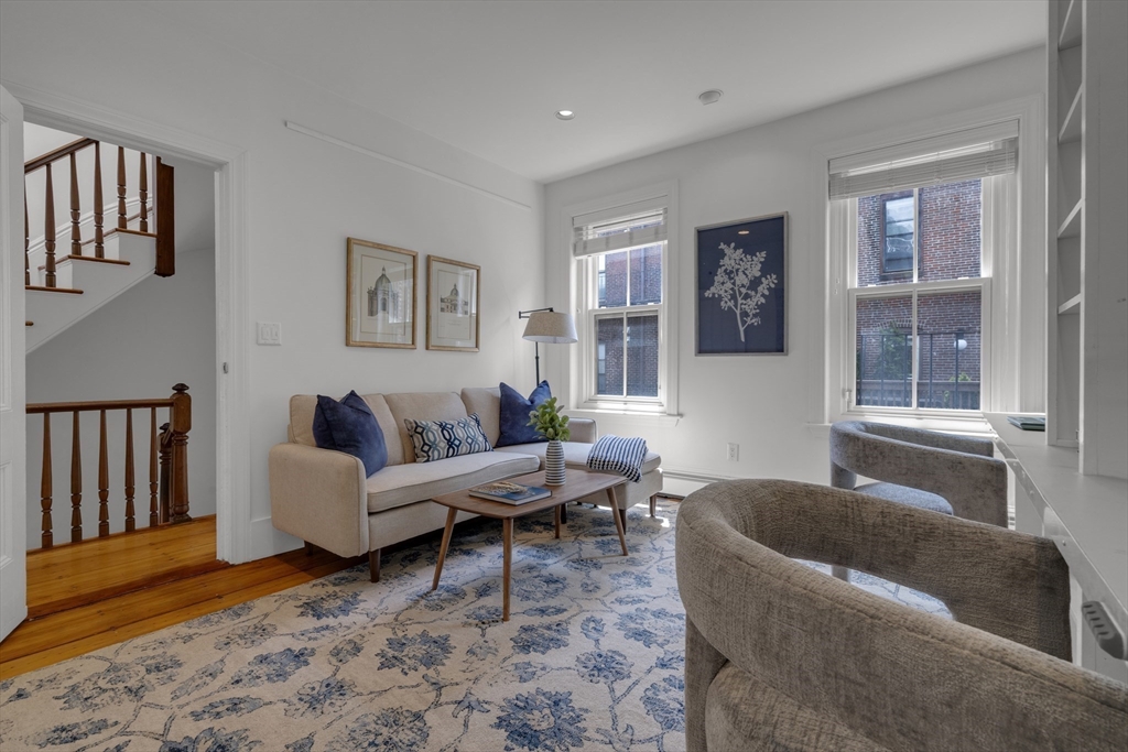 Living room with a beige sofa, blue floral rug, and tall windows overlooking brick buildings.