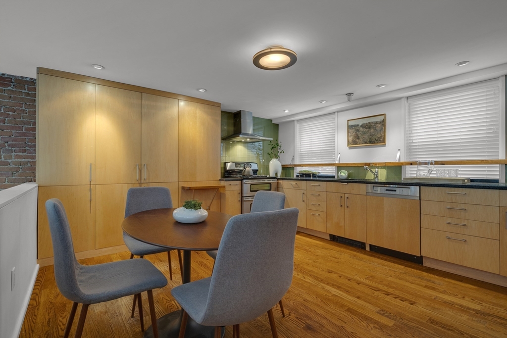 Kitchen with honey-colored wood cabinetry, hardwood floors, and a dining table beside an exposed brick wall.