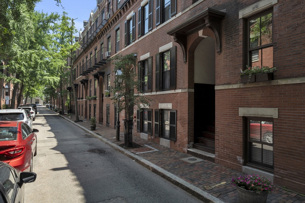 Red-brick row house with black shutters and a recessed entrance on a tree-lined street.