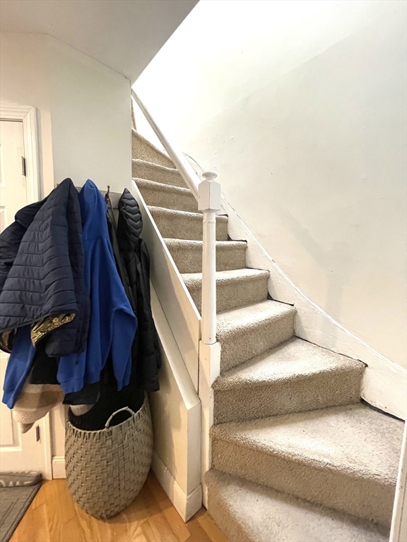 Carpeted staircase with a white railing, coats hanging beside it, and a woven basket below.