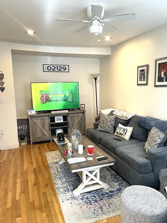 Living room with a gray sectional, ceiling fan, hardwood floors, and 02129 sign.