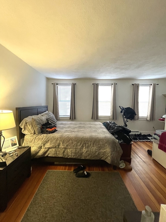 Furnished bedroom with a large bed, hardwood floors, and three curtained windows.