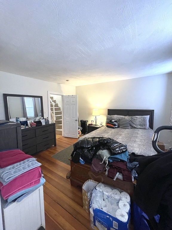 Bedroom with a large gray-covered bed, dark dresser, hardwood floors, and clothing piled across the bed.