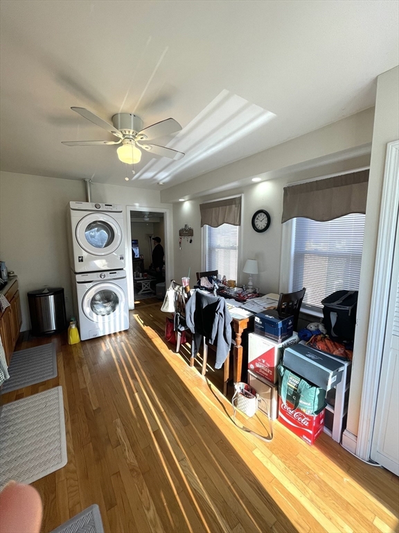 Cluttered room with stacked washer and dryer, a table, and sunlight across hardwood floors.