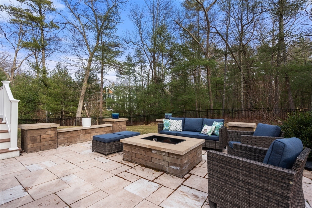 Stone patio with woven seating and a square fire pit, bordered by woods.