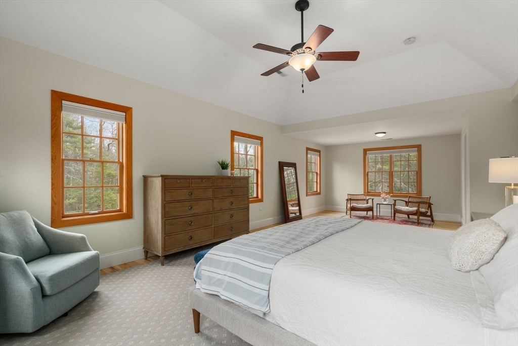 Staged bedroom with a large bed, wood-trimmed windows, ceiling fan, and blue armchair.