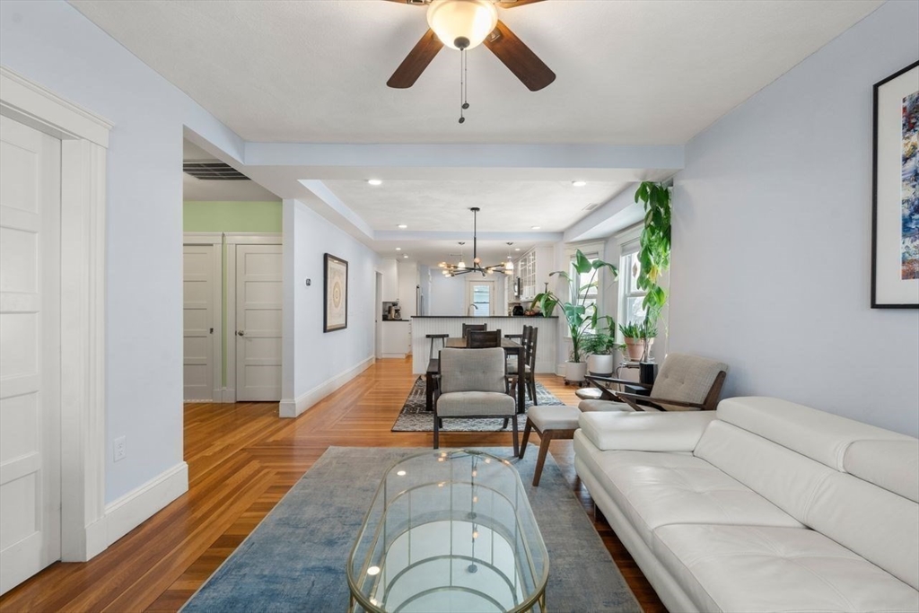 Living room with white sofa and glass coffee table, opening to a dining area and kitchen.