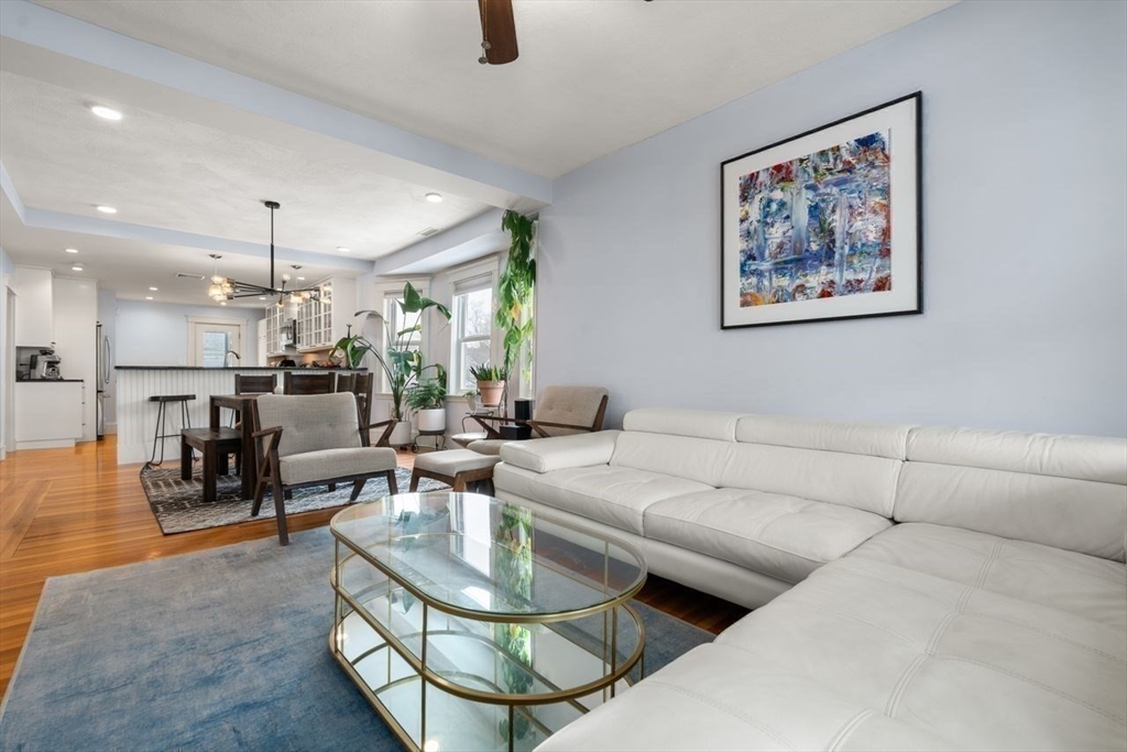 Open-plan living room with a white sectional, glass coffee table, and dining area beside the kitchen.