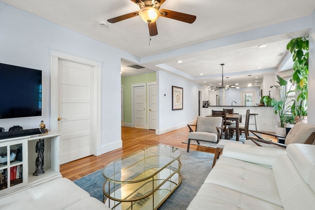 Open-plan living room with white sofas, a glass coffee table, hardwood floors, and kitchen dining area.