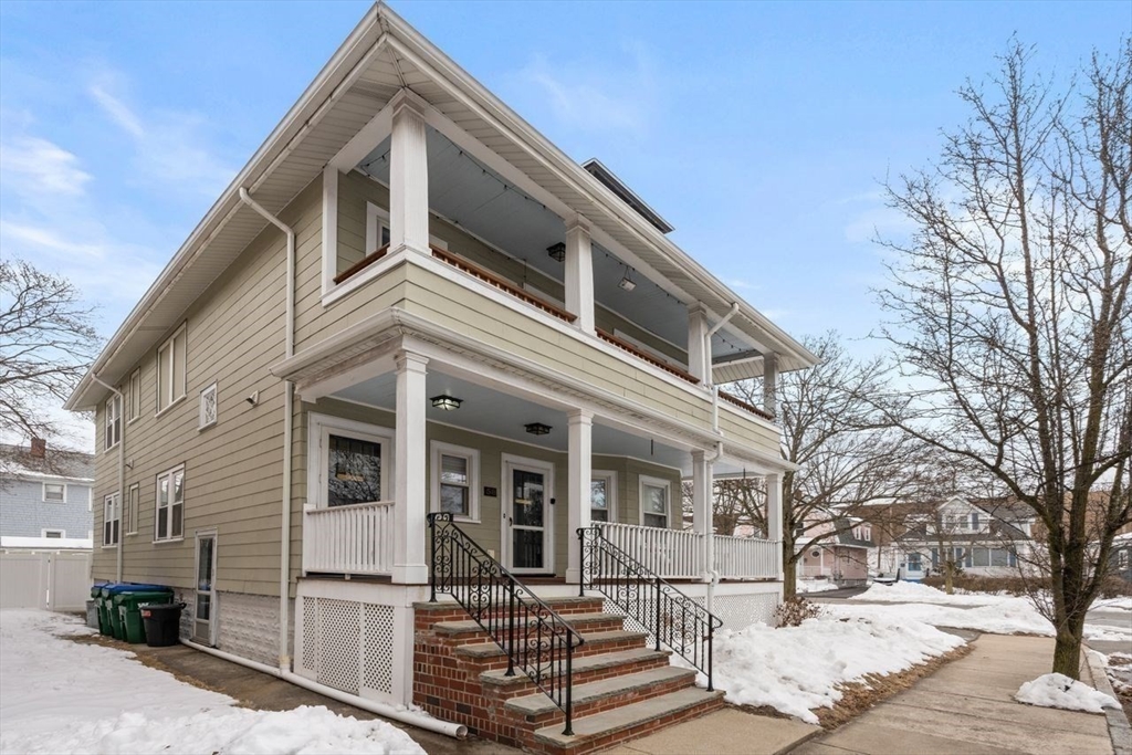 Beige two-story home with stacked covered porches and brick steps, photographed on a snowy day.