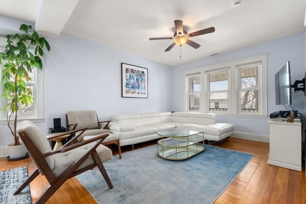 Living room with a white sectional, wooden lounge chairs, and large windows overlooking the street.
