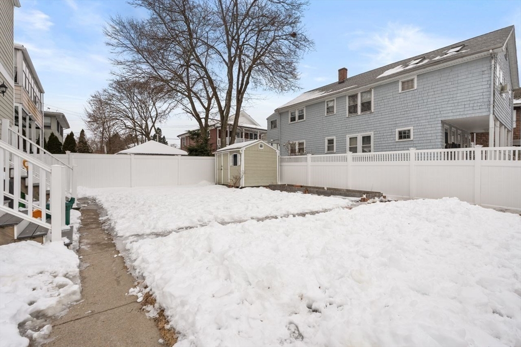 Snow-covered backyard enclosed by a white privacy fence, with a small shed and paved walkway.