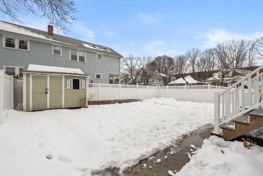 Snow-covered fenced backyard with a small shed beside the house and deck steps.