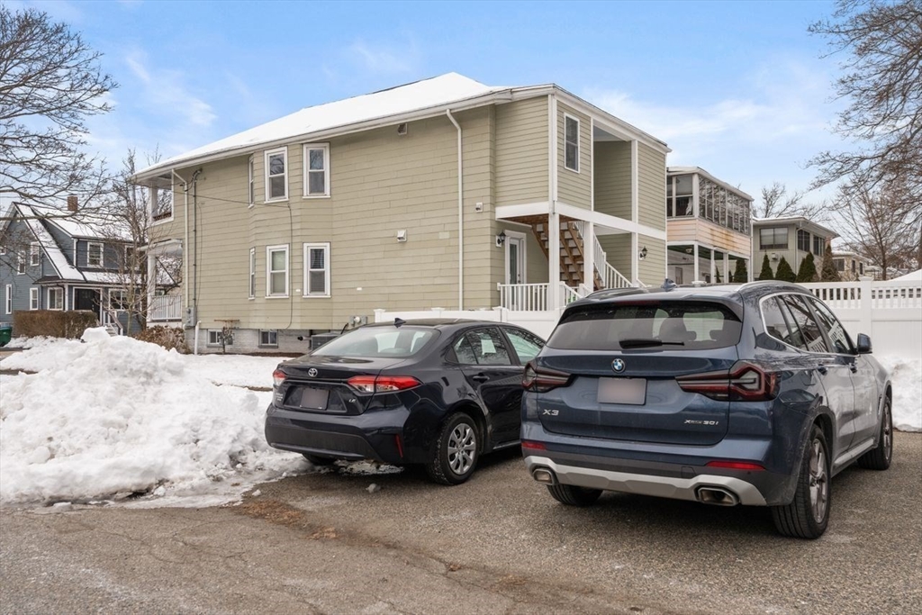 Beige two-story home with an elevated porch and exterior stairs beside a snowy parking area.