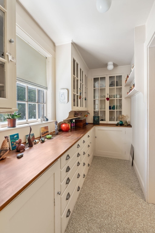 Kitchen pantry with cream cabinets, glass-front cupboards, and a long wooden countertop beside a window.