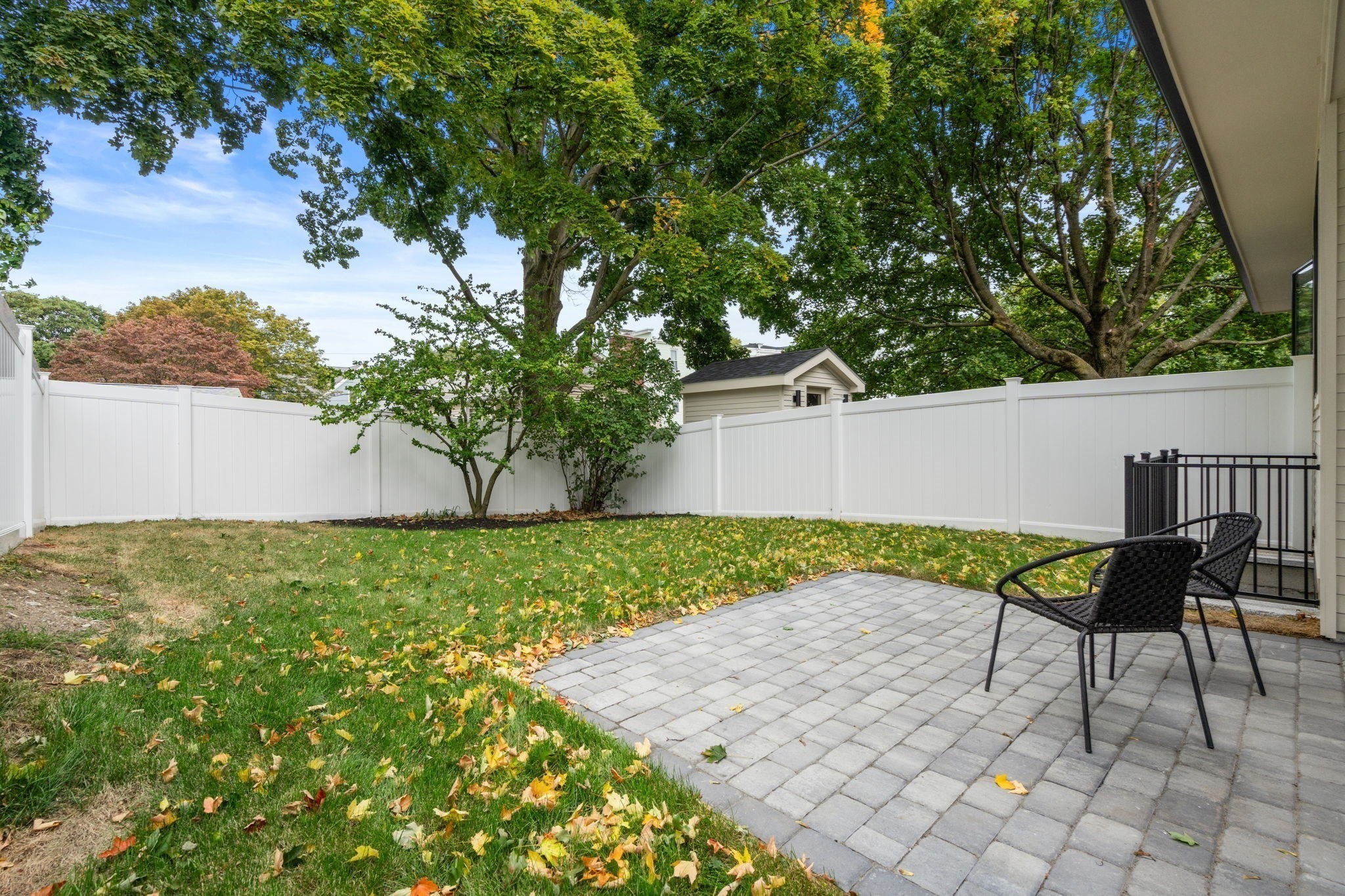 Fenced backyard patio with gray pavers and black chairs beside a leaf-strewn lawn.