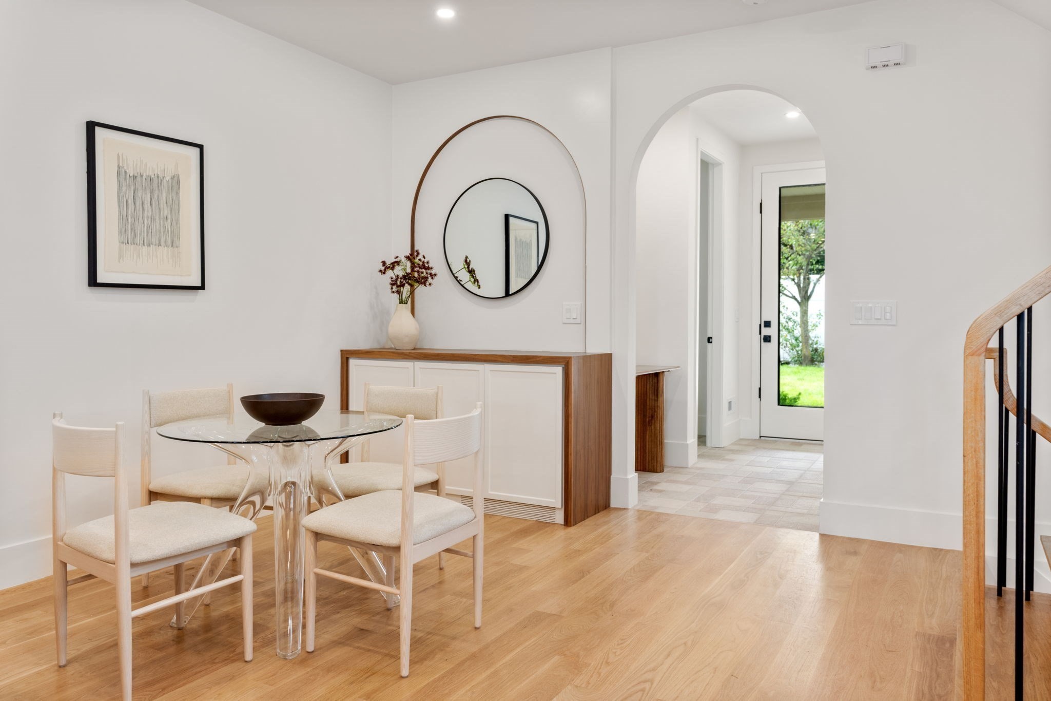 Dining area with a glass table, pale upholstered chairs, arched mirror, and built-in cabinet.