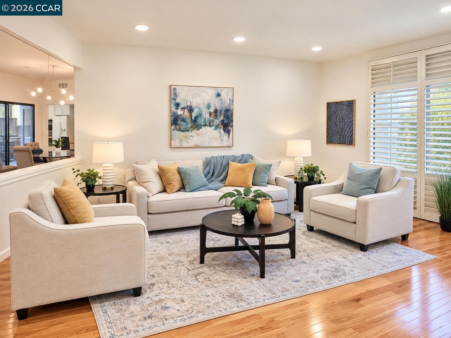 Living room with cream seating, blue and ochre pillows, plantation shutters, and hardwood floors.