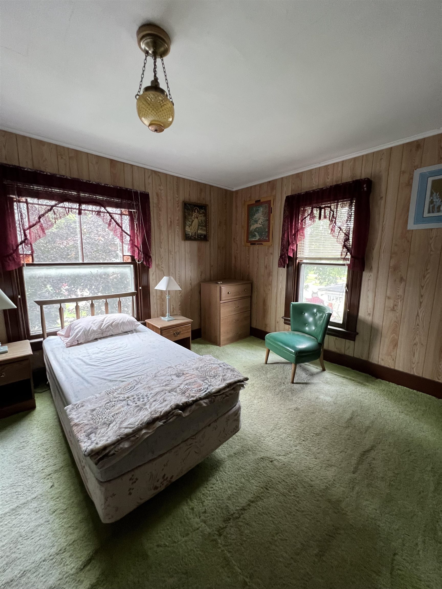 Bedroom with a single bed, green carpet, wood paneling, and a green chair by two curtained windows.
