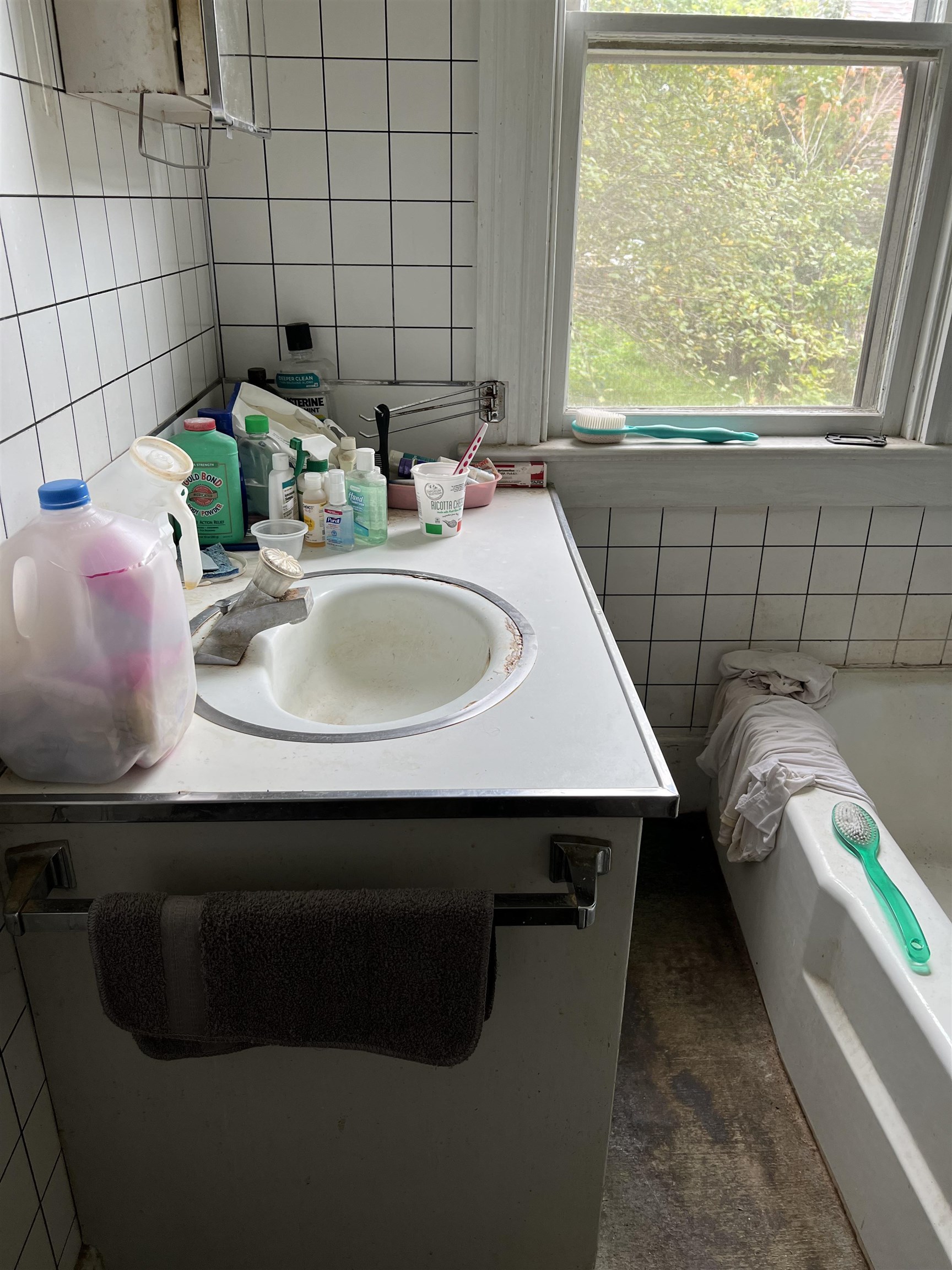 Bathroom with a tiled sink vanity and bathtub; toiletries crowd the counter beside a window.