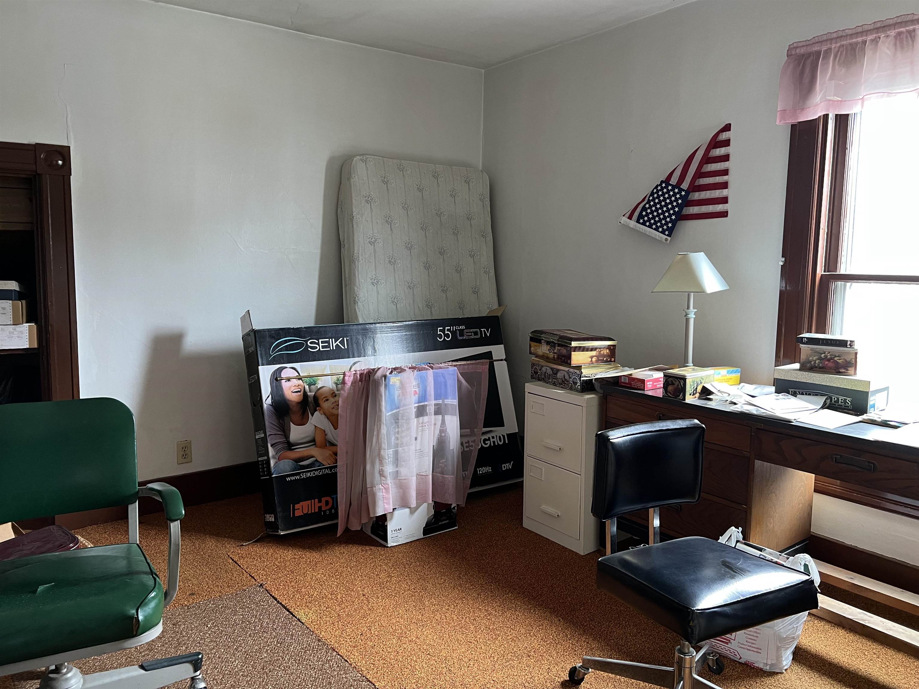 Cluttered room with a desk, filing cabinet, chairs, and a mattress leaning against the wall.