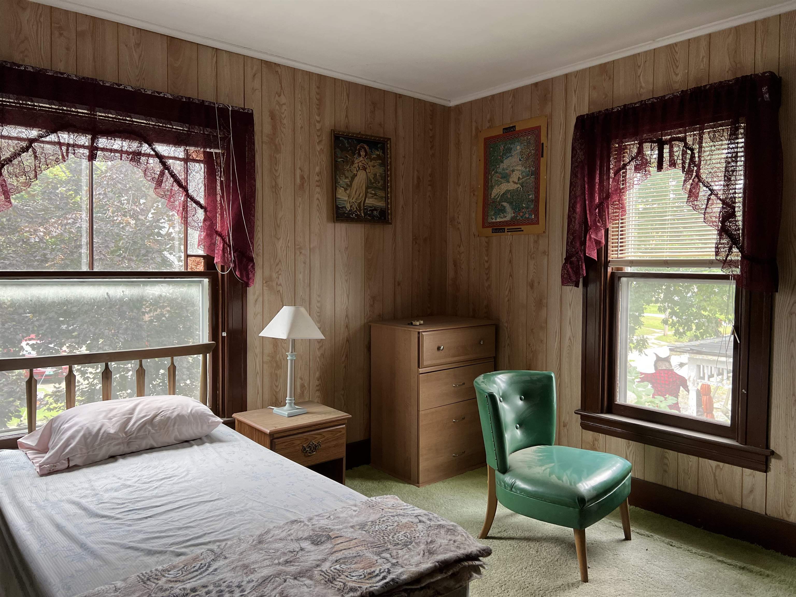 Bedroom with wood-paneled walls, burgundy curtains, and a green chair beside the bed.