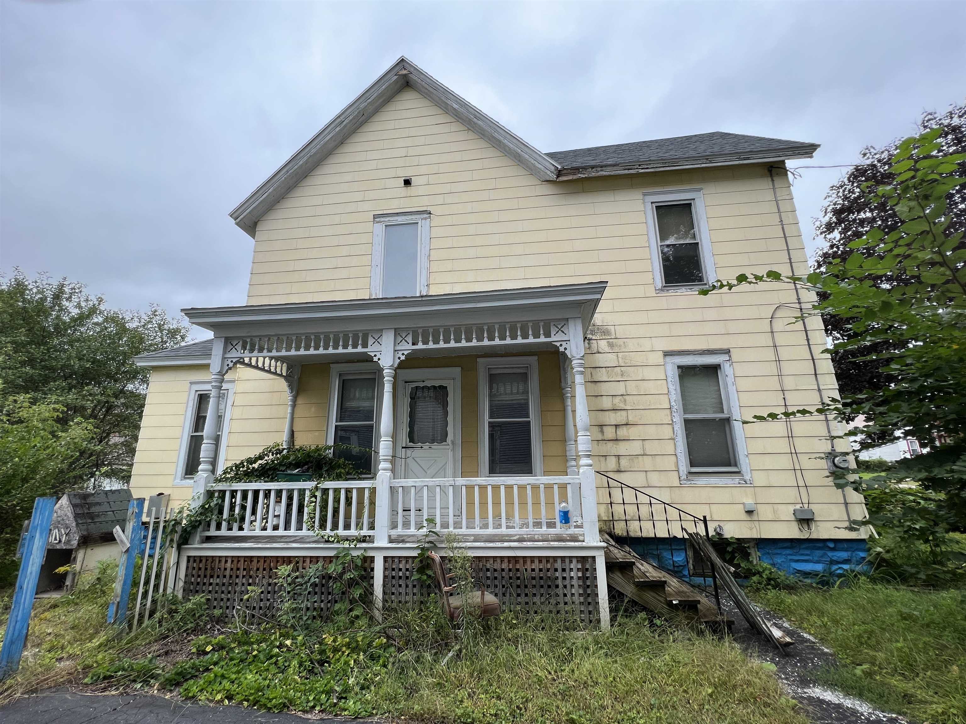 Two-story yellow house with a decorative white porch and overgrown yard.