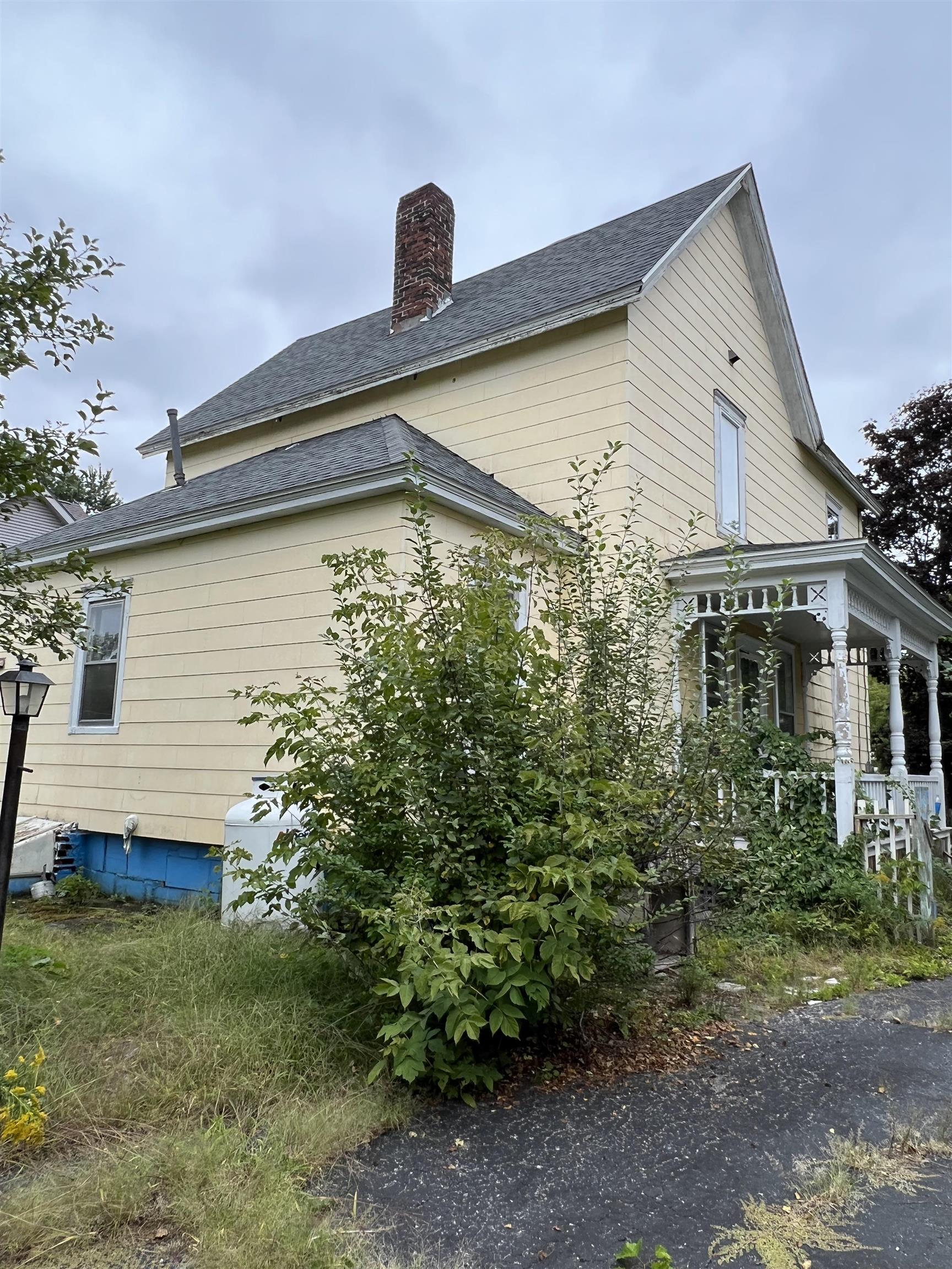 Yellow two-story house with a steep shingle roof, brick chimney, and shrub-lined covered porch.